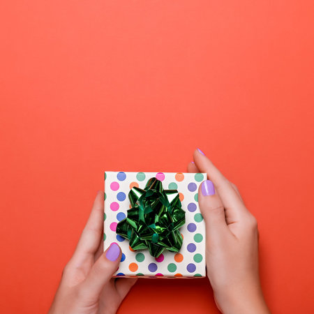 Woman manicured hands holding green giftbox on orange background with confetti, copy space, top...