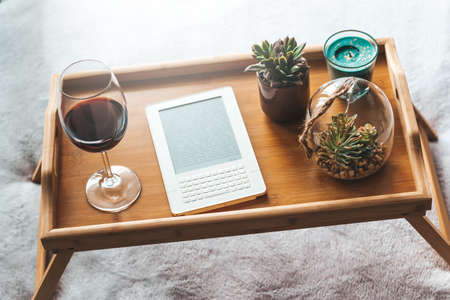 Electronic book, glass of wine, house plants on wooden tray. Reading and relaxing during self isolation at home as preventative measure against virus outbreak. What to do during quarantine concept.の写真素材