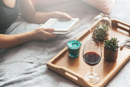 Electronic book, glass of wine, house plants on wooden tray. Reading and relaxing during self isolation at home as preventative measure against virus outbreak. What to do during quarantine concept.の写真素材