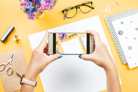 Woman taking photo of her office desk. Note books, accessories, stationery, flowers on table top, woman workspaceの写真素材