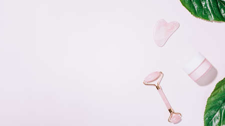 Facial roller, gua sha and moisturizing cream on green leaves. Natural cosmetics and facial tools for massage on pink background. Rose quartz beauty roller, heart shape gua sha flat lay.の写真素材