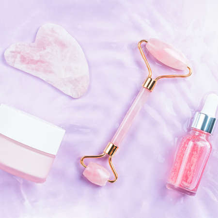 Pink hyaluronic acid serum, rose quartz roller, gua sha and face cream in water, top view. Anti aging cosmetics in pink water, top viewの写真素材