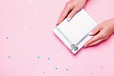 Woman hands holding silver gift box on pastel pink background with confetti, top view, copy space.