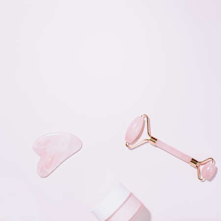 Facial roller, gua sha and moisturizing cream on green leaves. Natural cosmetics and facial tools for massage on pink background. Rose quartz beauty roller, heart shape gua sha flat lay.の写真素材