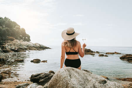Woman in bikini and hat alone at beach holding glass of wine and enjoying peaceful scenery, back view. Woman enjoying time at mediterranean beach, sunbathing and drinking wineの写真素材