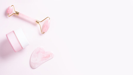 Pink quartz facial roller, gua sha and moisturizing cream on pastel pink. Tools for skin care...