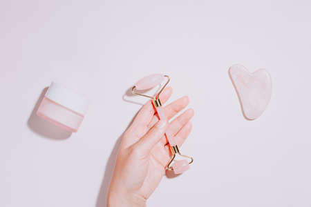 Facial roller, gua sha and moisturizing cream with green leaves. Natural cosmetics and facial tools on pink. Rose quartz beauty roller, heart gua sha flat lay. Tools for skin care routines, top viewの写真素材