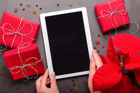 Tablet, red gift boxes, poinsettia flower, chocolate star and Christmas lights on dark gray tabletopの写真素材