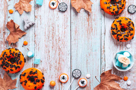 Orange Halloween doughnuts decorated with chocolate bats, licorice lollipops and spooky marshmallows on blue wooden table. Halloween frame, flat lay, copy spaceの写真素材