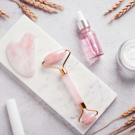 Facial roller, gua sha, serum and moisturizing cream on marble background. Facial tools with wheat decor on white. Rose quartz beauty roller, gua sha flat lay. Tools for skin care routines, top viewの写真素材