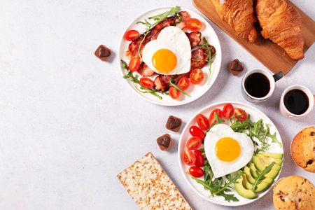 Fried eggs in heart shape, bacon, avocado and vegetables on two white plates, coffee cups and muffins- couple breakfast on Valentines Day or honeymoon on gray background, food border, copy spaceの写真素材