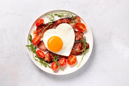 Fried eggs in heart shape, bacon and vegetables on white plate - surprise breakfast for husband or boyfriend on Valentines Day or Fathers Day. Men's Breakfast, minimal styleの写真素材