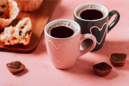 Two coffee cups with heart in pink and black colors, muffins and chocolates closeupの写真素材