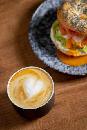 Heart latte art coffee scene with a bagel with egg, avocado, and fish on a wooden table. Perfect breakfast or brunchの写真素材