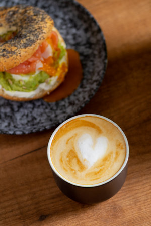Heart latte art in a cozy coffee scene with a bagel topped with egg, avocado, and fish on a wooden table. Perfect breakfast or brunch ambiance.の写真素材