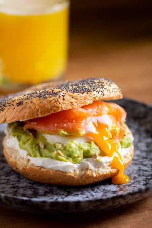 Bagel with poached eggs, cream cheese, avocado, and fish. A cozy and nutritious breakfast or brunch arrangement on a wooden table with orange juiceの写真素材