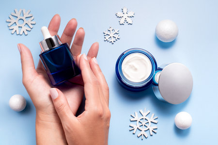 Winter skincare beauty concept with woman hands holding face serum and cream moisturizer on a blue backgroundの写真素材