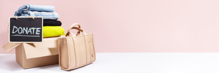 Box filled with clothes and handbag, with a Donate sign on a blackboard, against a pink backgroundの写真素材