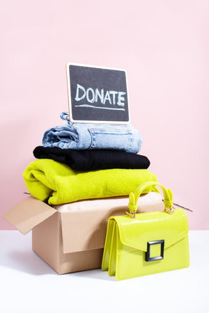 A cardboard box filled with folded clothes and a yellow handbag, with a Donate sign on a blackboard, against a pink background, minimal style still lifeの写真素材