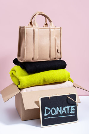 A cardboard box filled with folded clothes and handbag, with a Donate sign on a blackboard, against a pink background, minimal style still lifeの写真素材
