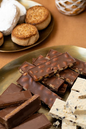 A detailed close-up of traditional Spanish holiday treats, chocolate turron with whole almonds, creamy nougat, and Polvorones on golden plates, Christmas or winter holiday setting.の写真素材