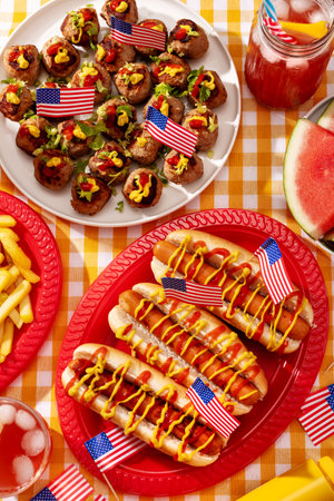 Top view of patriotic American food on a checkered yellow table with hot dogs, fries, meatballs, watermelon, and drinks decorated with USA flags.の写真素材
