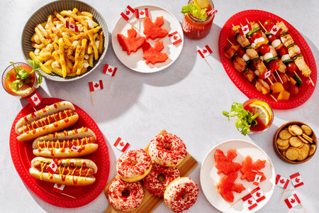 Top view frame of Canada Day party food with poutine, hot dogs, fruit, skewers, and donuts decorated with Canadian flags on a light background.の写真素材
