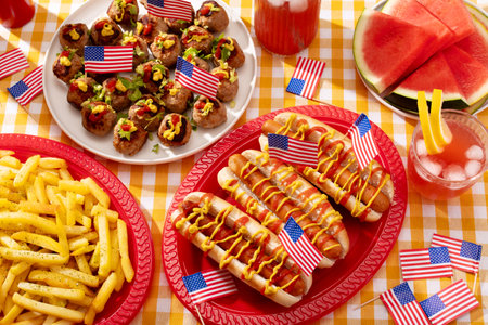 Top view of patriotic American food on a checkered yellow table with hot dogs, fries, meatballs, watermelon, and drinks decorated with USA flags.の写真素材