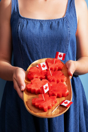 Woman in a denim dress holding a wooden tray with maple leaf shaped watermelon slices, each topped with a small Canadian flag, festive treat for Canada Day.の写真素材