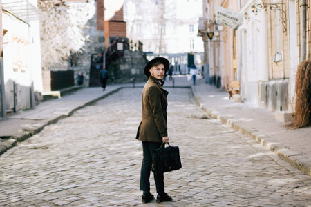 handsome young bearded hipster man guy in hat Fedora on street with suitcase. Retro vintage fashion...
