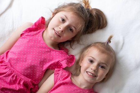 Two happy sisters in pink dresses are lying on the bed and smilingの写真素材