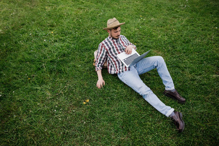 Young man sitting on green grass and working on laptop outdoors