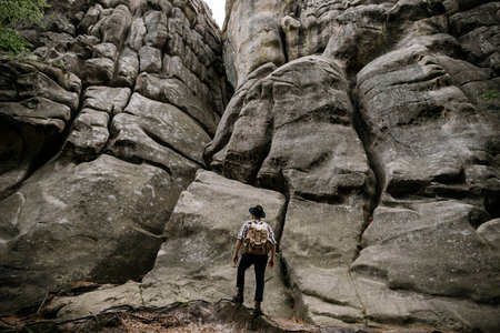 A hipster traveler stands with a backpack in front of a large rock in the mountains