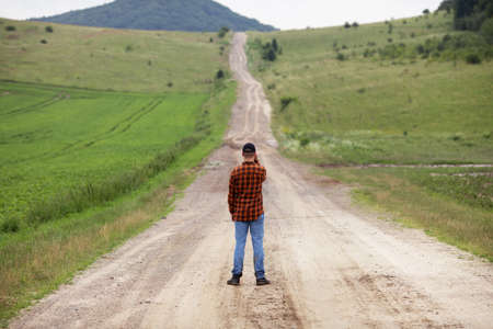 A man in a plaid shirt and blue jeans stands in the middle of a field road in a rural areaの写真素材