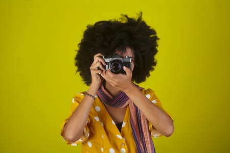 Beautiful indian girl with afro curly hairstyle talking pictures with retro camera on yellow backgroundの写真素材