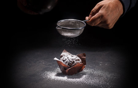 View of the hand of the pastry chef putting powdered sugar on delicious chocolate muffin on dark...