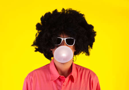 Curly man wearing sunglasses blowing bubble with chewing gum on yellow backgroundの写真素材