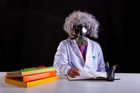 Crazy science teacher in white coat with unkempt hair sitting at the desk wears a gas maskの写真素材