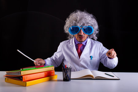 Crazy science teacher in white coat with unkempt hair in funny eye glasses sitting at the deskの写真素材
