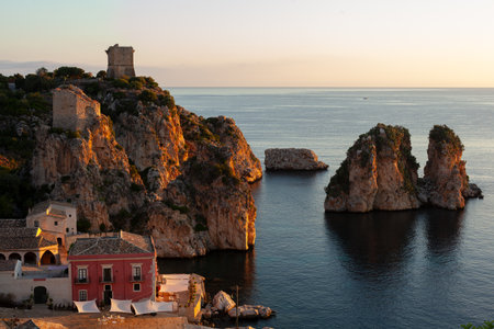 A beautiful beach with a small red building at sunrise. The water is calm and the sky is a beautiful blue. Tonnara di Scopello, province of Trapani, Sicily. Italyの写真素材