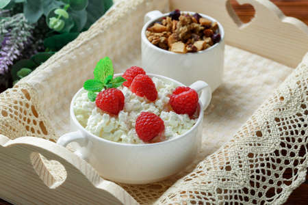 Homemade breakfast: cottage cheese with fresh raspberries and granola. In a white porcelain bowl, on a wooden trayの写真素材