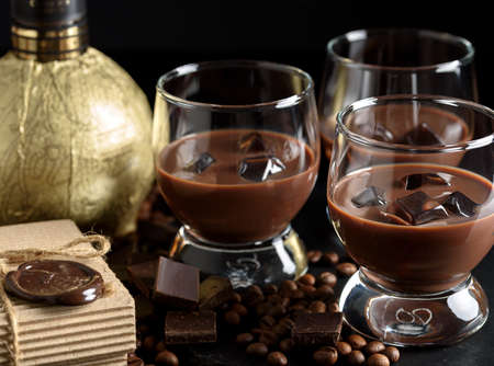 Chocolate, coffee liqueur in glass glasses with ice cubes. With coffee beans, chocolate, cinnamon and a bottle of liqueur in gold foil. On a black backgroundの写真素材