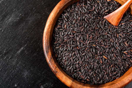 Isolated handful of raw black (nero) rice in the wood bowl on black stone background.の写真素材