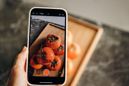 Woman taking photo of persimmon fruits on smartphone in kitchen.