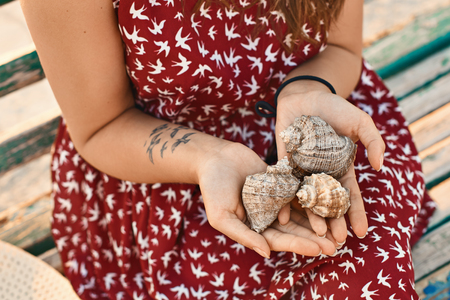 Young woman wearing a red dress with a bird pattern and a tattoo is holding three beautiful seashells with her hands over the lap while sitting on the wooden benchの写真素材