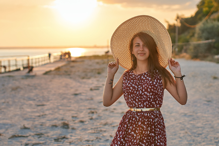 Fashion portrait of a young beautiful girl in a dress with bird pattern holding a straw beach hat and being happy in the rays of sunset sunlight at the summer seasideの写真素材