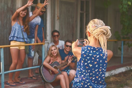 Young blonde girl with dreads takes a photo of a groups of her friends with her smartphone near the wooden holiday cabinの写真素材