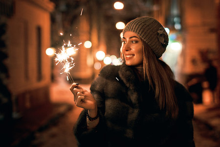 Beautiful young woman in fur coat holding a sparkler enjoys winter Christmas mood in old snowy...