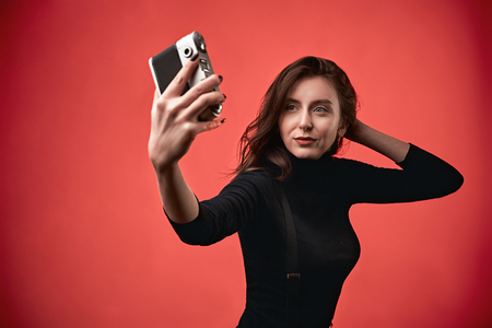 Portrait of a stylish young brunette girl making a selfie photograph shot with a vintage film photo camera on coral red backgroundの写真素材