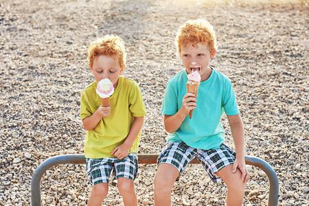Two cute red curly boys brothers are happy while getting dirty and eating ice cream on the summer beachの写真素材